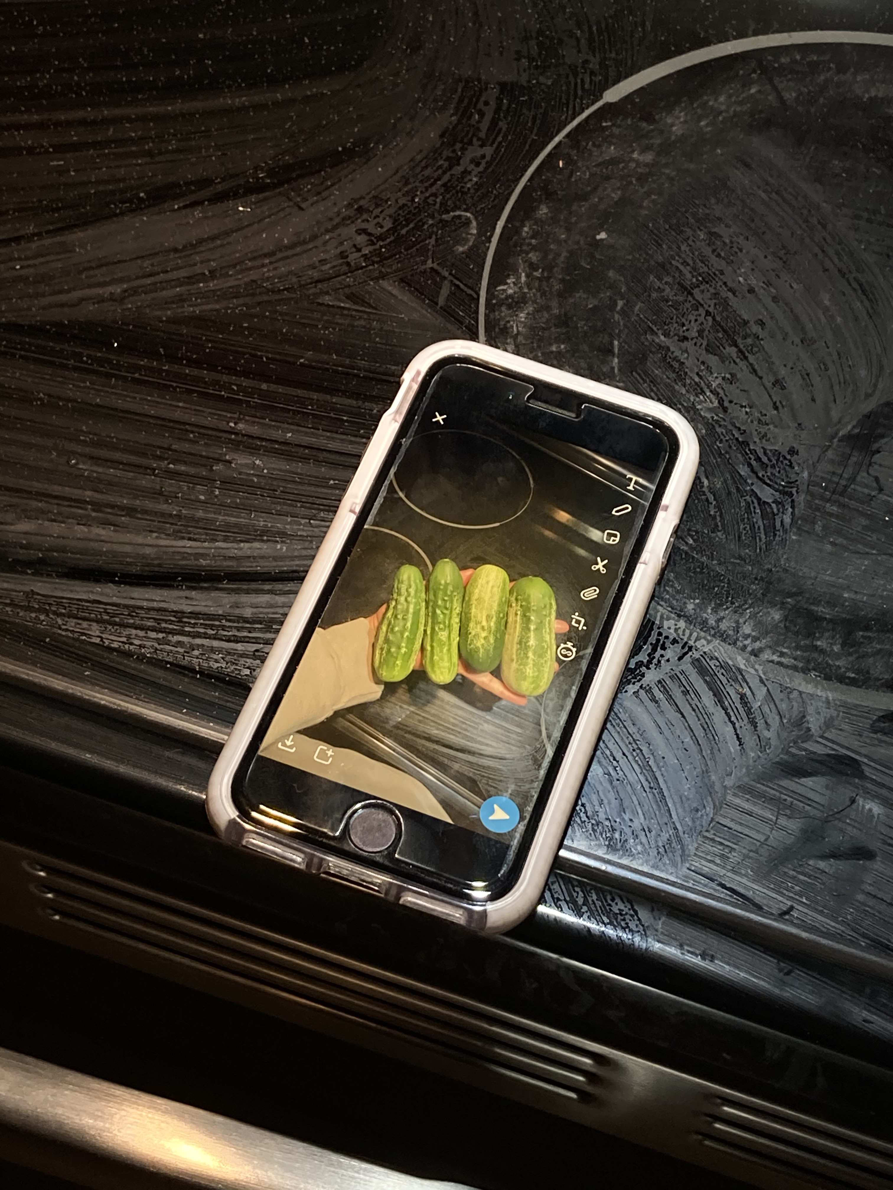 a phone sits on a stovetop, and on the phone is a picture of a hand holding 4 squat cucumbers